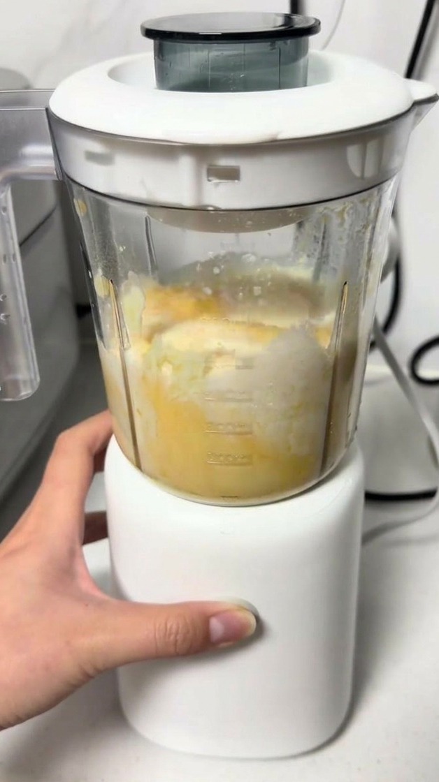 Ręka obsługująca mały, biały blender, w którym miksowana jest żółta masa serowo-jajeczna.