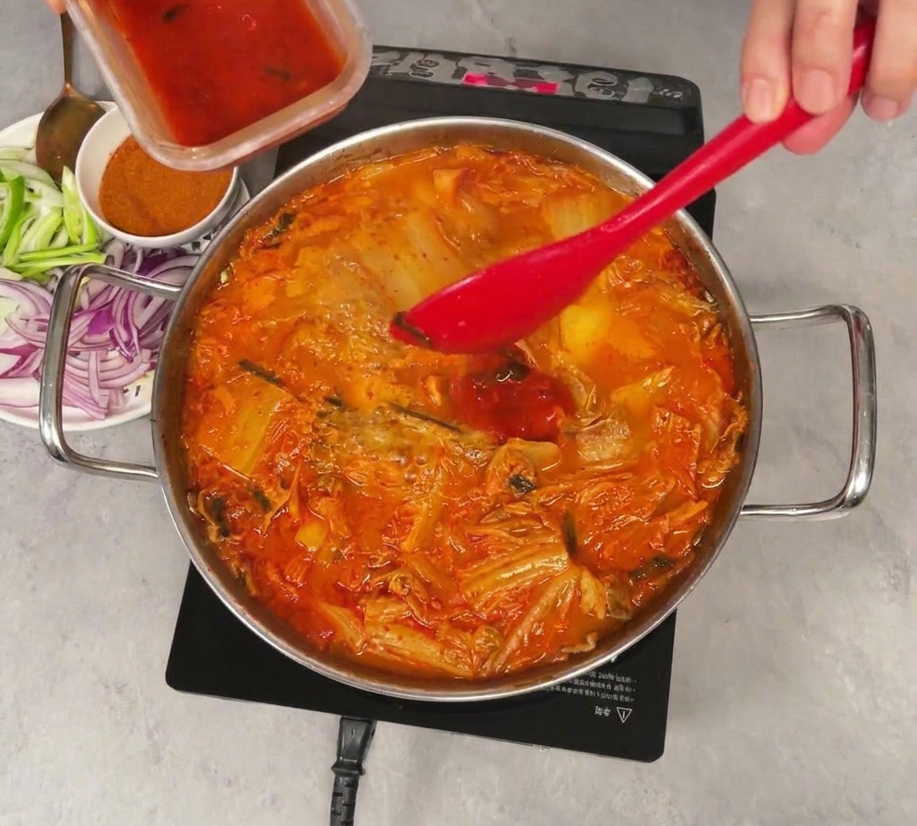 Jasnoczerwony sok z kimchi wlewany z małego plastikowego pojemnika do gotującego się garnka z gulaszem, mieszanego czerwoną szpatułką.