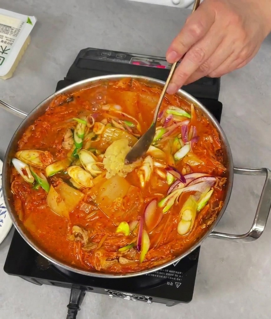 Dodawanie łyżki świeżo posiekanego czosnku do bulgoczącej zupy kimchi na patelni.