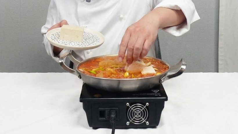 Kucharz delikatnie kładący kwadratowe plastry naturalnego tofu do bulgoczącego garnka z czerwoną zupą kimchi.