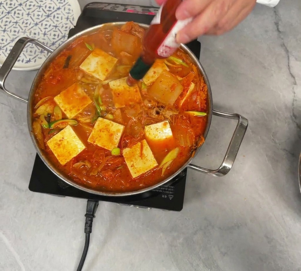 Dodawanie ostrego sosu Tabasco z małej buteleczki do gotującego się garnka czerwonej zupy kimchi na której ułożono tofu.