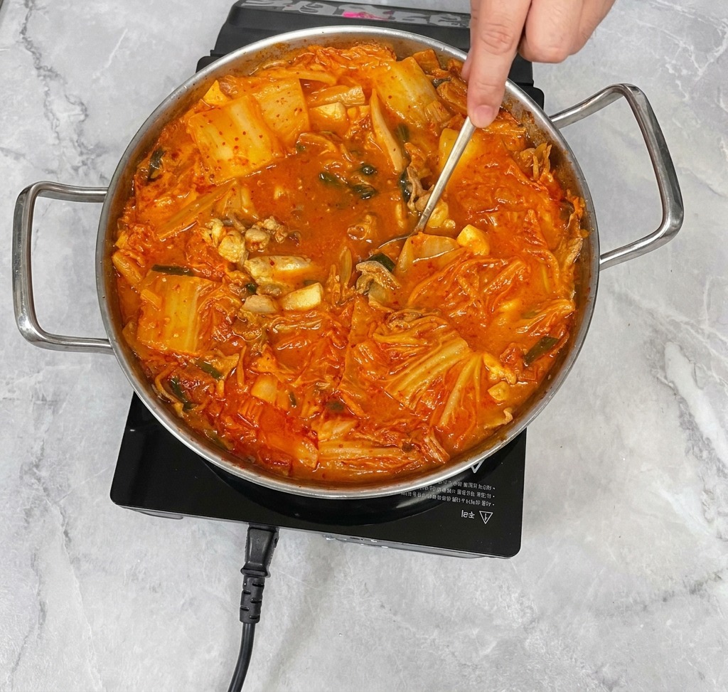 Ręka używająca łyżki ze stali nierdzewnej do delikatnego mieszania intensywnie czerwonej zupy kimchi, łącząc przyprawy z bulionem.