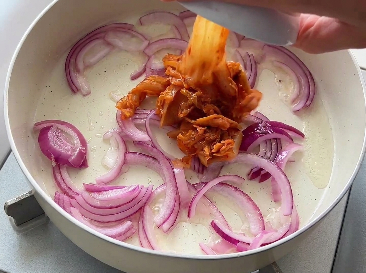 Posiekane kimchi dodawane z małej miski na patelnię z podsmażoną czerwoną cebulą.