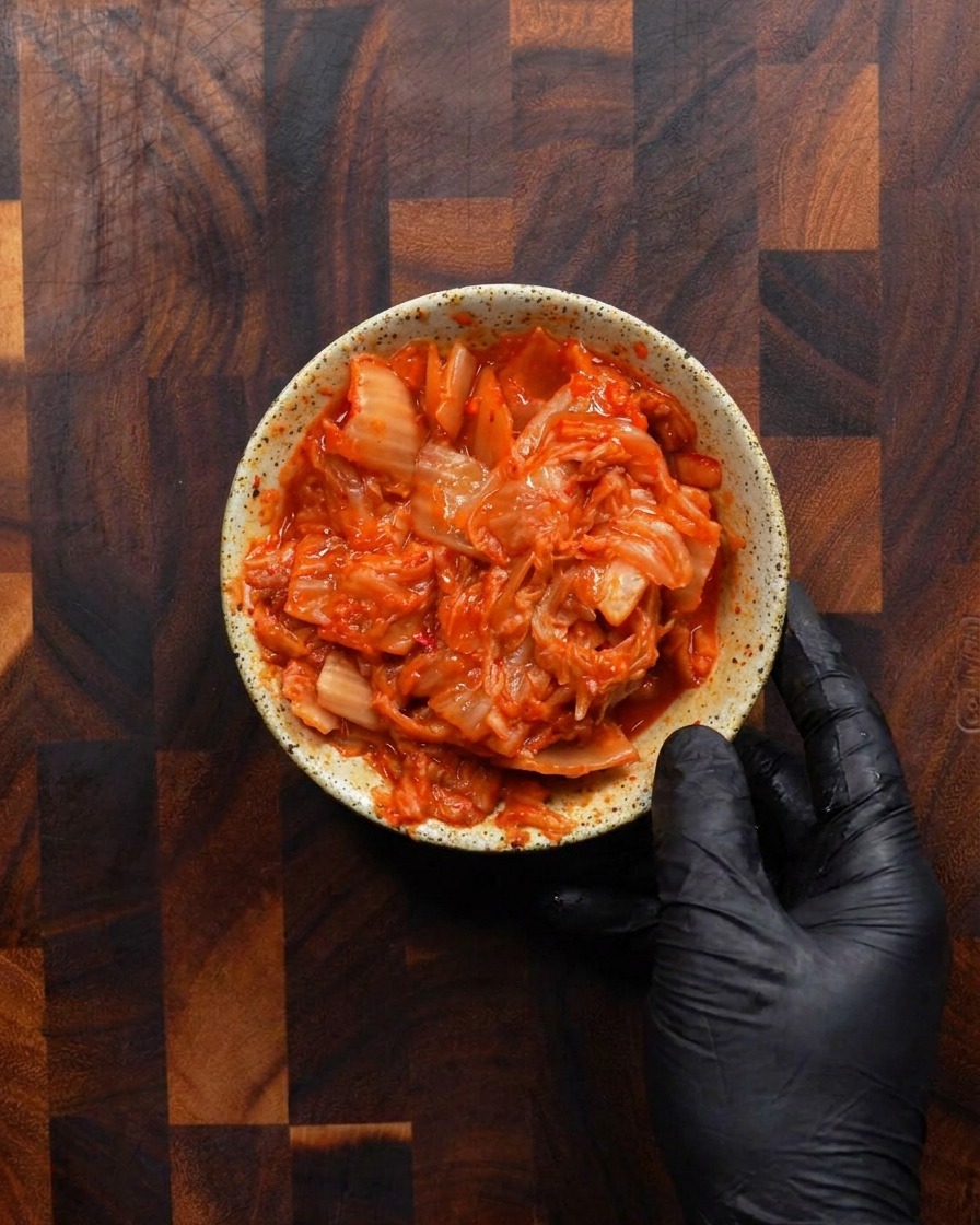 Dłoń w rękawiczce trzymająca małą, rustykalną, nakrapianą miseczkę wypełnioną po brzegi jaskrawoczerwonym, posiekanym sfermentowanym kimchi.
