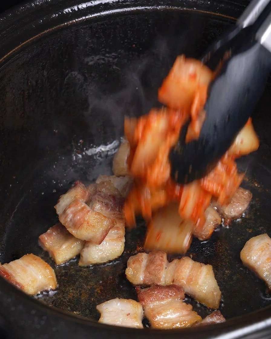 Posiekane czerwone kimchi dodawane szczypcami do zrumienionego boczku w czarnym garnku.