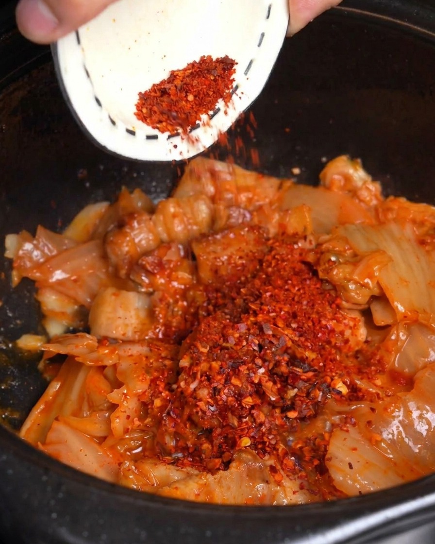 Czerwone sproszkowane chili zsypywane z małej miseczki na podsmażone kimchi i boczek w czarnym garnku.