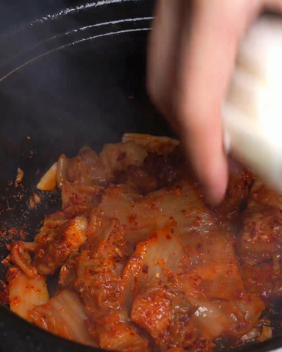 Dłoń wrzucająca grube plastry świeżej białej cebuli do gorącego, parującego garnka z jasnoczerwonym podsmażonym kimchi i boczkiem.