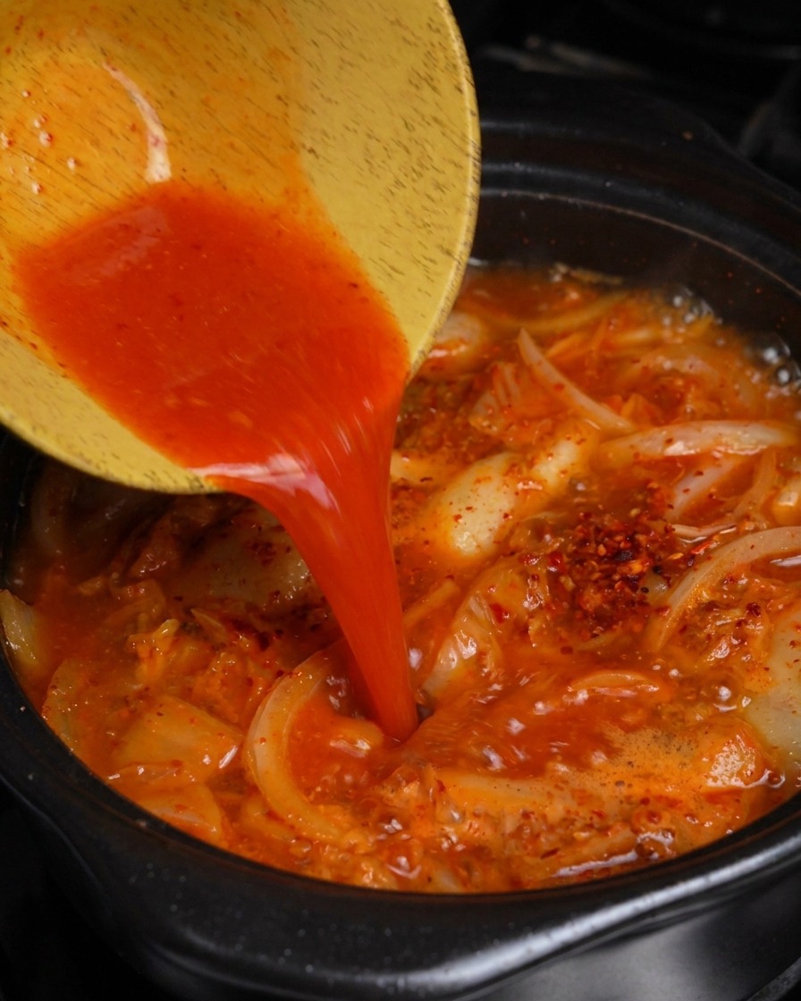 Wlewanie jaskrawoczerwonego soku z kimchi z drewnianej miski do garnka z bulgoczącym gulaszem wieprzowym z kimchi.