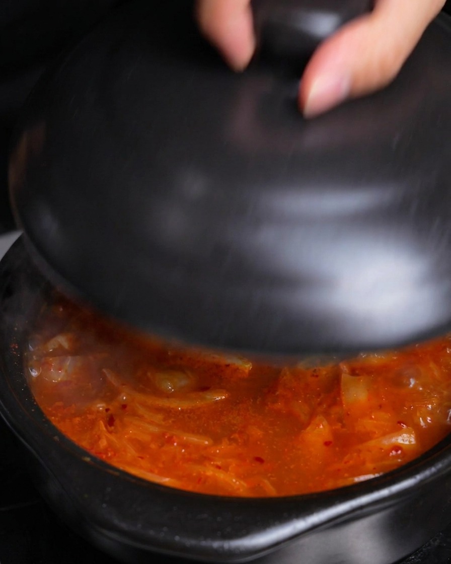 Dłoń kładąca czarną, ceramiczną pokrywkę na garnku z gorącym, bulgoczącym gulaszem kimchi, aby pozwolić mu się dusić.