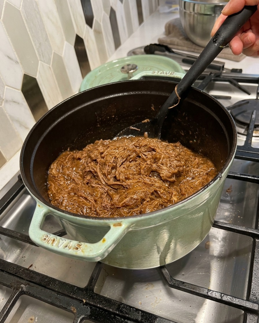 Szarpana wołowina mieszana z bogatym, ciemnym sosem barbecue w zielonym garnku za pomocą czarnej łopatki.