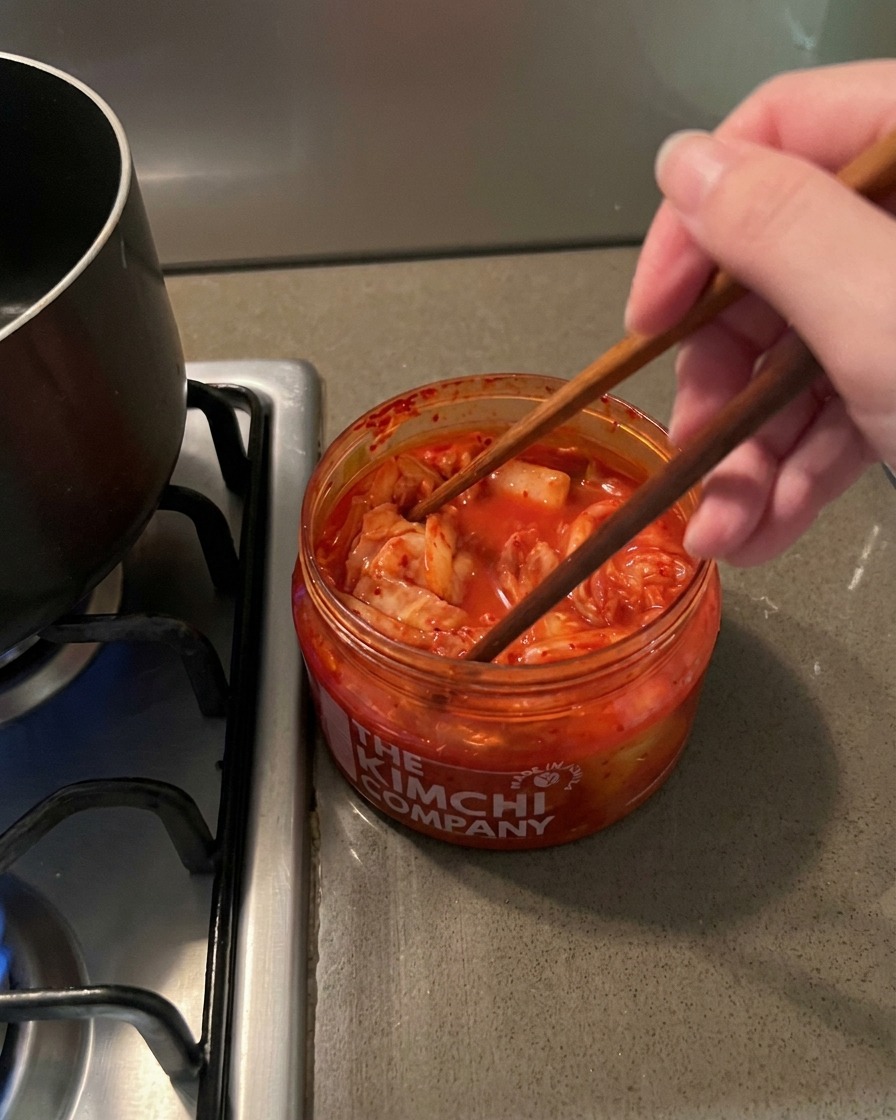 Drewniane pałeczki wyjmujące porcję czerwonego, pikantnego kimchi z przezroczystego słoika.