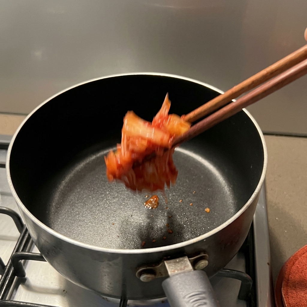 Jasnoczerwone kimchi wrzucane pałeczkami do ciemnego rondla na kuchence.