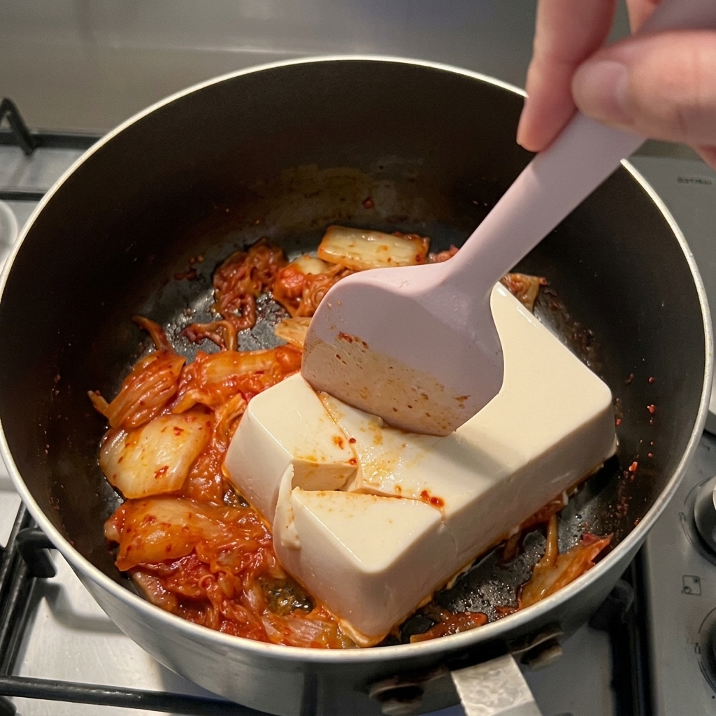 Różowa silikonowa szpatułka przecinająca blok białego tofu w rondlu z czerwonym kimchi.