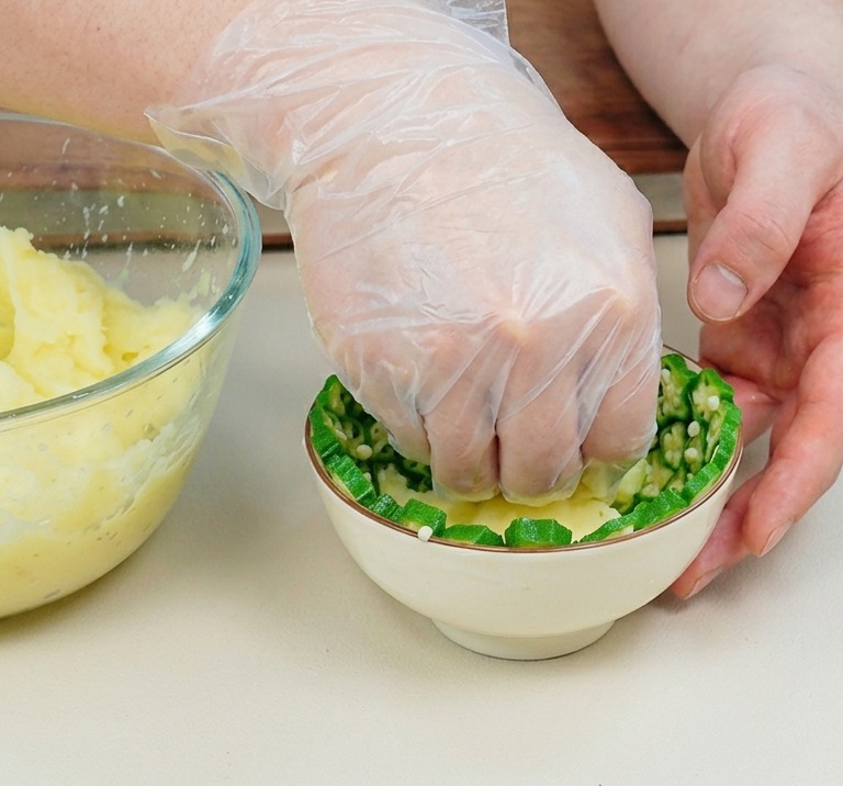 Dłoń w rękawiczce dociska purée ziemniaczane do miseczki wyłożonej plasterkami okry.