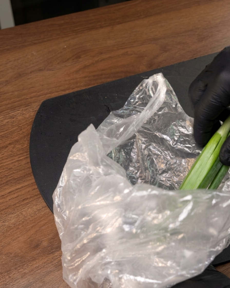Ręka w rękawiczce wyjmująca świeże zielone warzywa z plastikowej torebki na ciemnym blacie.
