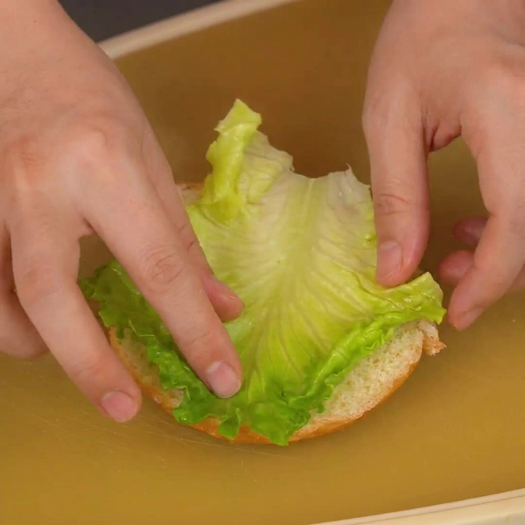 Dłonie kładące liść zielonej sałaty na dolnej części bułki do burgera.