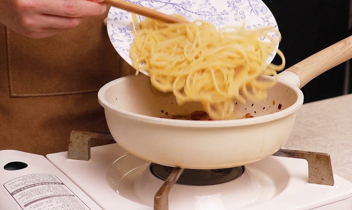 Zsuwanie ugotowanego spaghetti z niebiesko-białego w kwiatowe wzory talerza na białą patelnię z chrupiącym guanciale.