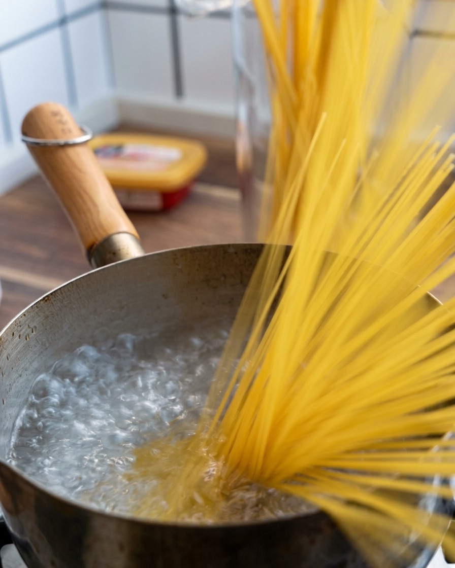Garść suchego makaronu spaghetti wrzucana do garnka z intensywnie gotującą się wodą.