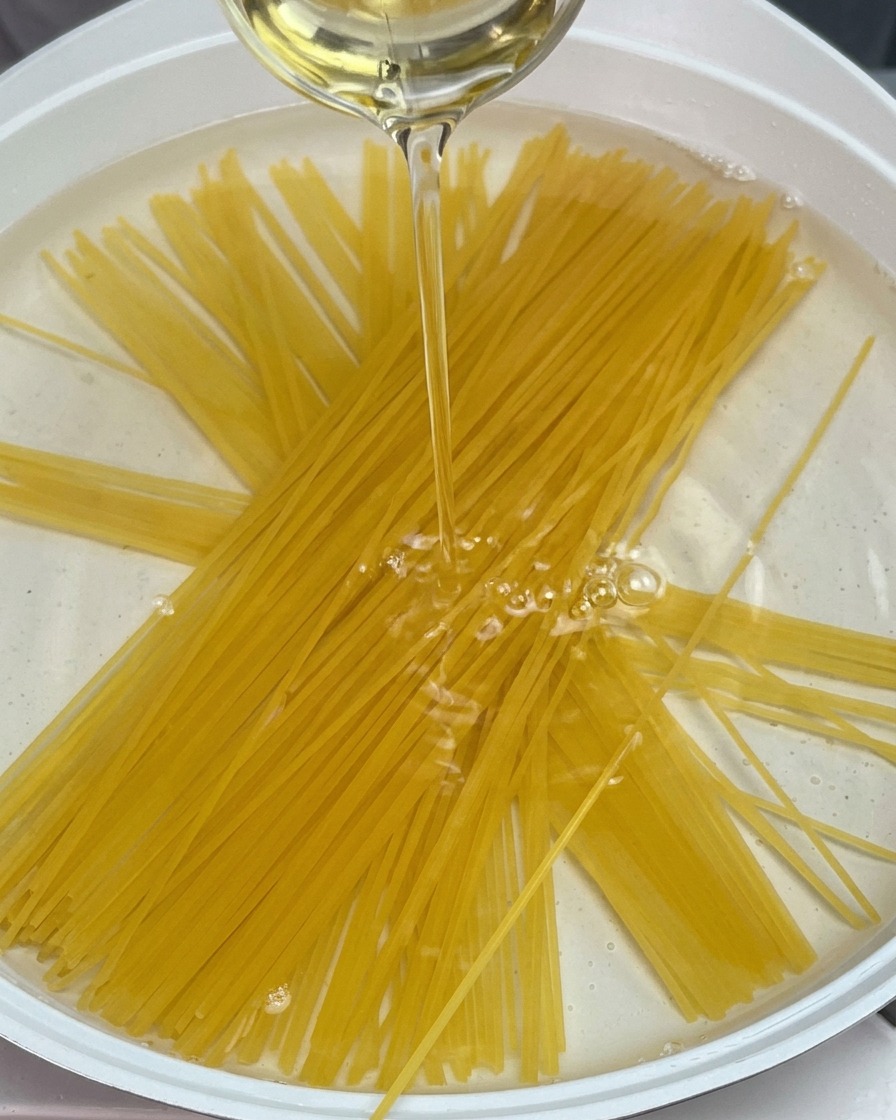 Surowy makaron spaghetti zanurzony w garnku z wodą, do której wlewany jest strumień oleju.