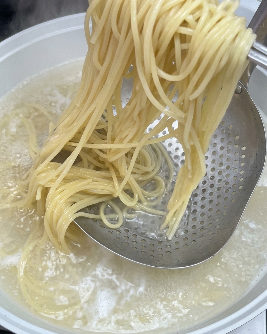 Ugotowany makaron spaghetti wyjmowany z garnka z wrzącą wodą za pomocą metalowej łyżki cedzakowej.