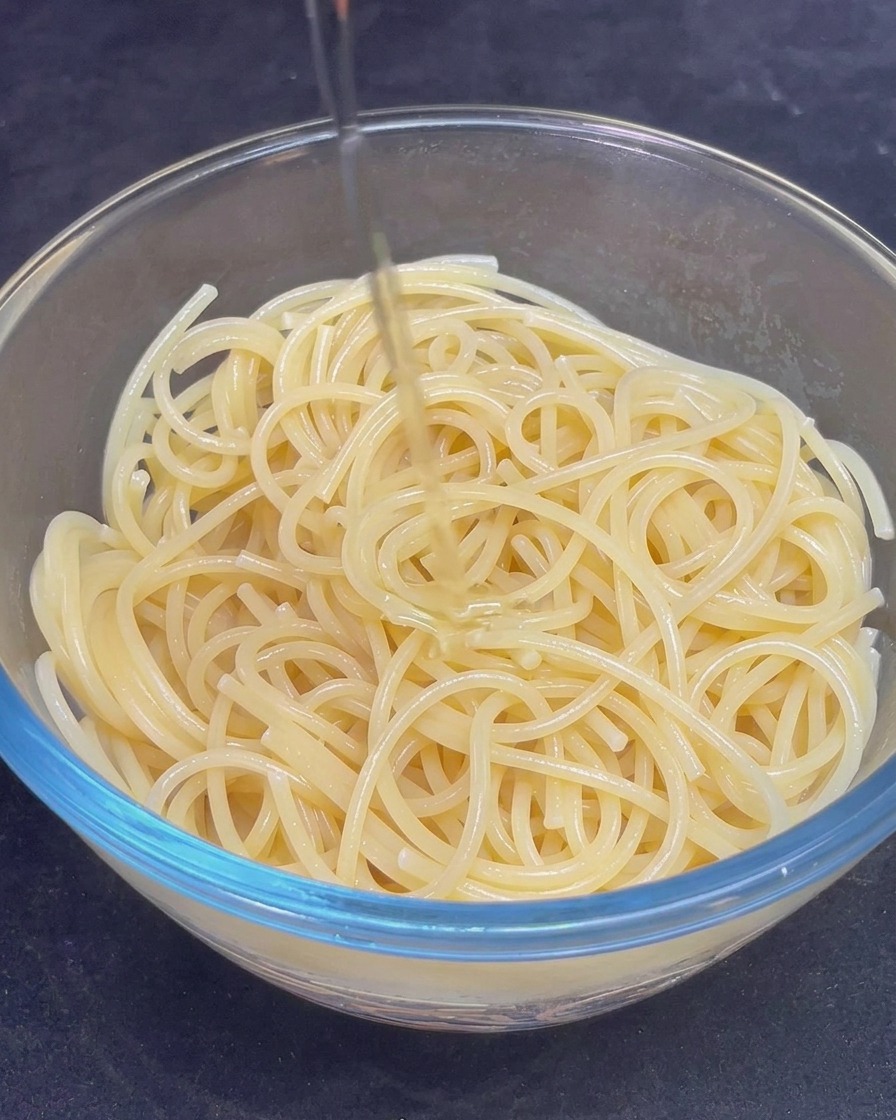 Strumień złocistej oliwy z oliwek wlewany do szklanej miski wypełnionej ugotowanym makaronem spaghetti.
