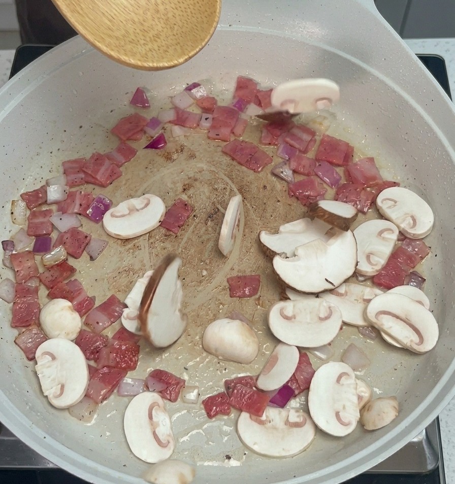 Plasterki pieczarek i grzybów shiitake wrzucane na patelnię do boczku i cebuli za pomocą drewnianej łyżki.