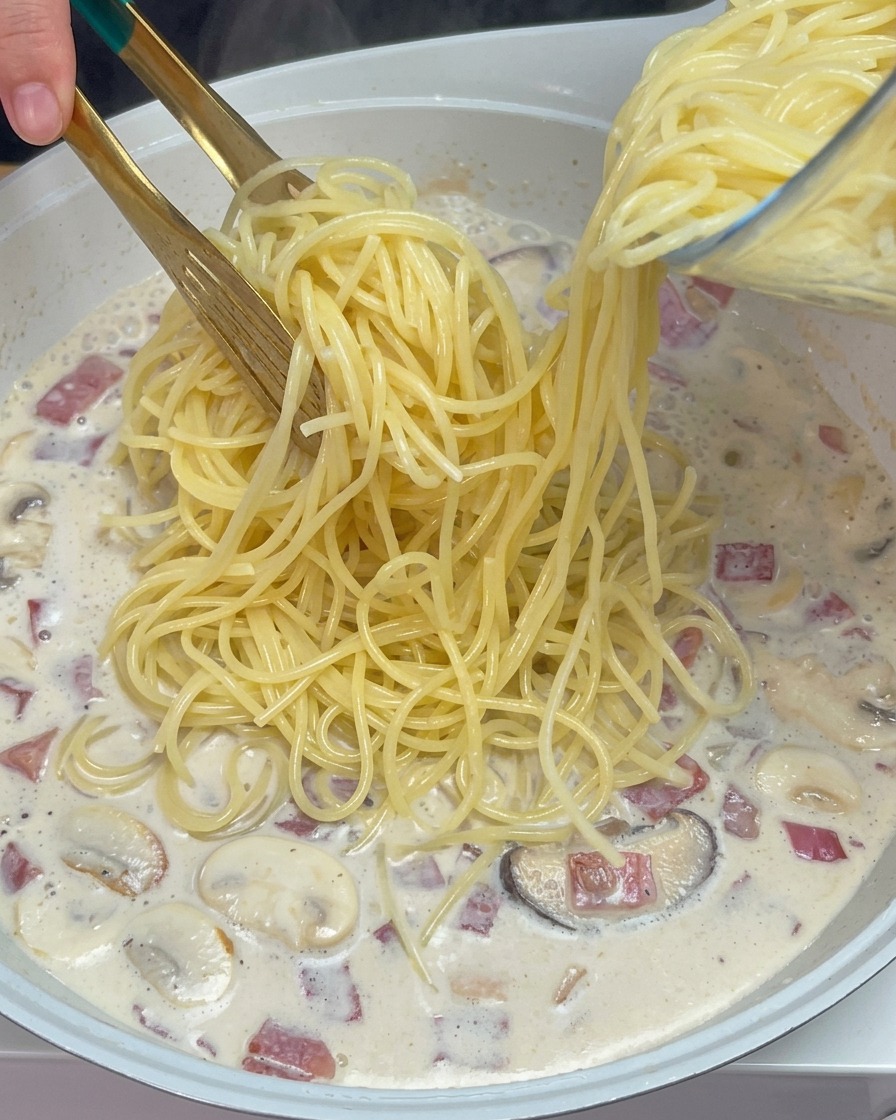 Szczypce nakładające ugotowane spaghetti do gotującego się kremowego sosu z grzybami i boczkiem.