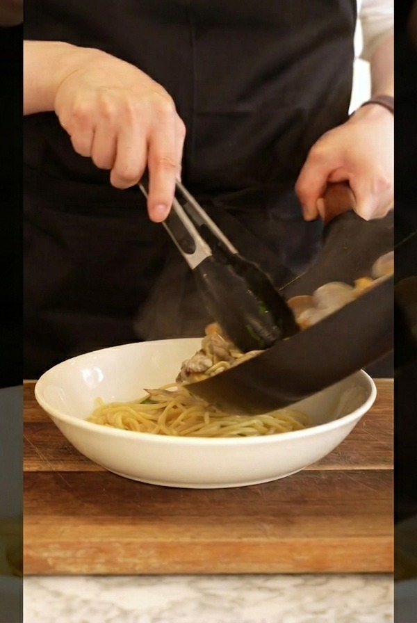 Kucharz używający szczypiec do przekładania gorącego spaghetti z małżami do białej miski.