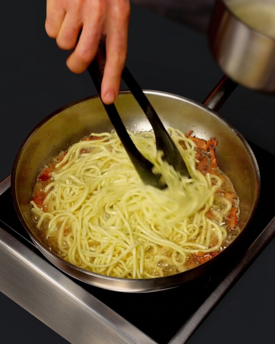 Mały rondel dolewający gorącą wodę po makaronie na patelnię ze spaghetti i guanciale podczas mieszania szczypcami.