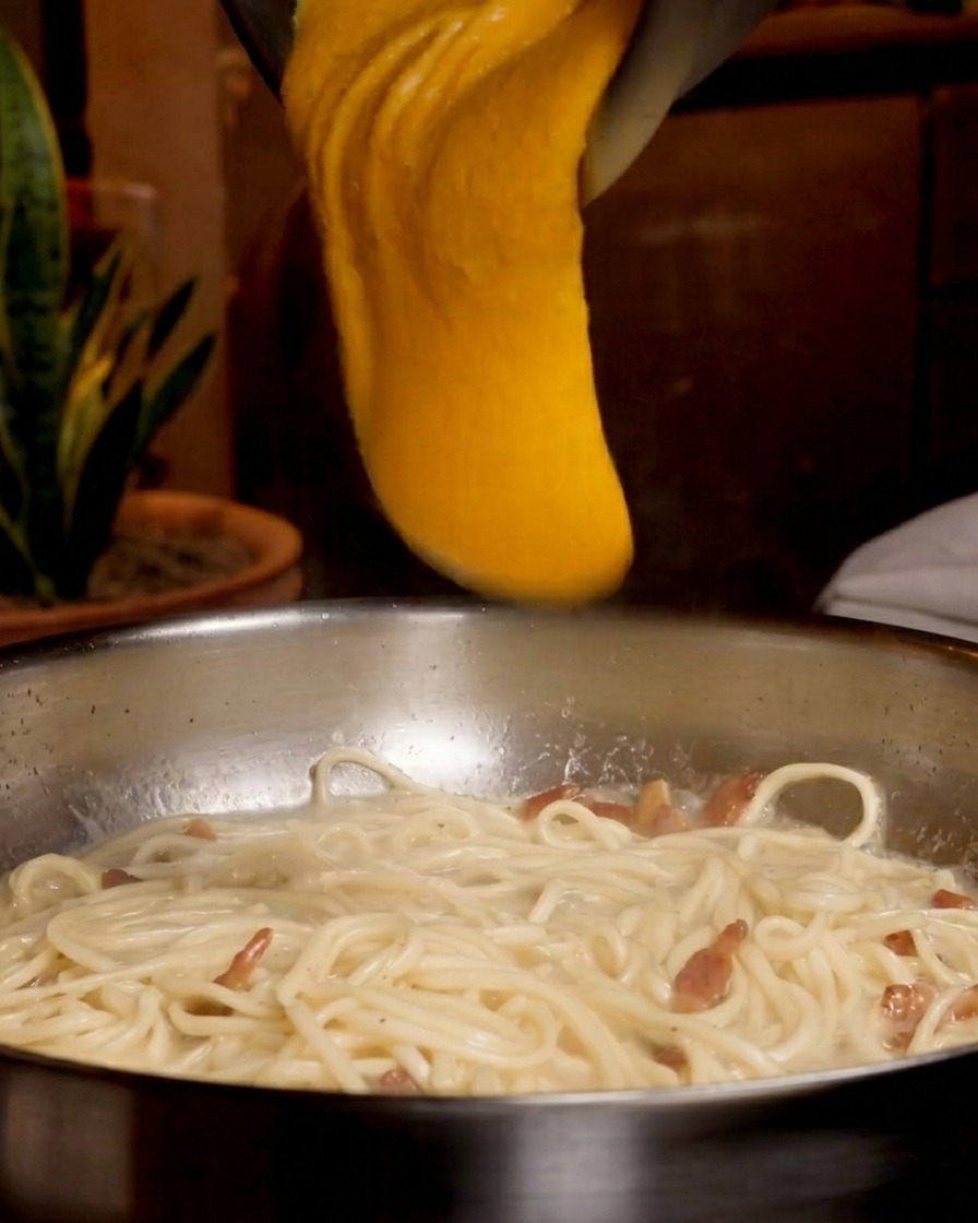 Gęsta, jaskrawopomarańczowa mieszanka jajek i sera jest wylewana z metalowej miski na patelnię ze spaghetti i guanciale.