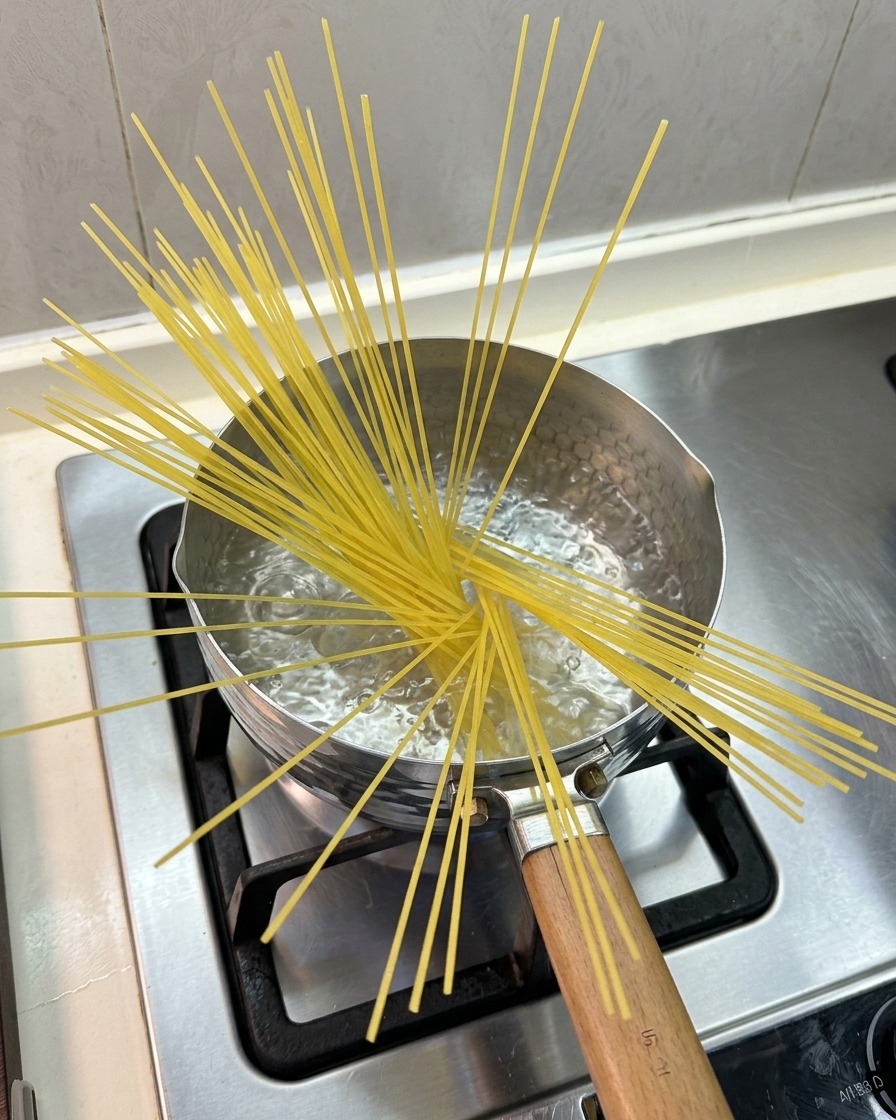 Surowy makaron spaghetti wystający z gotującej się wody w niewielkim garnku na kuchence gazowej.
