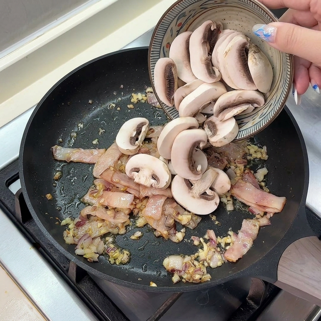 Pokrojone pieczarki wsypywane z ceramicznej miski na gorącą patelnię z boczkiem i cebulą.
