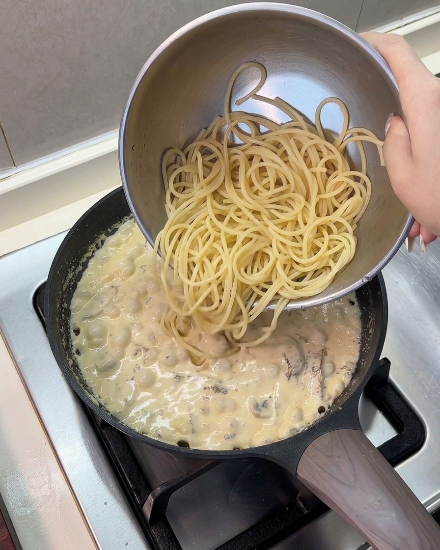 Ugotowany makaron spaghetti przekładany ze stalowej miski na patelnię z gotującym się sosem.