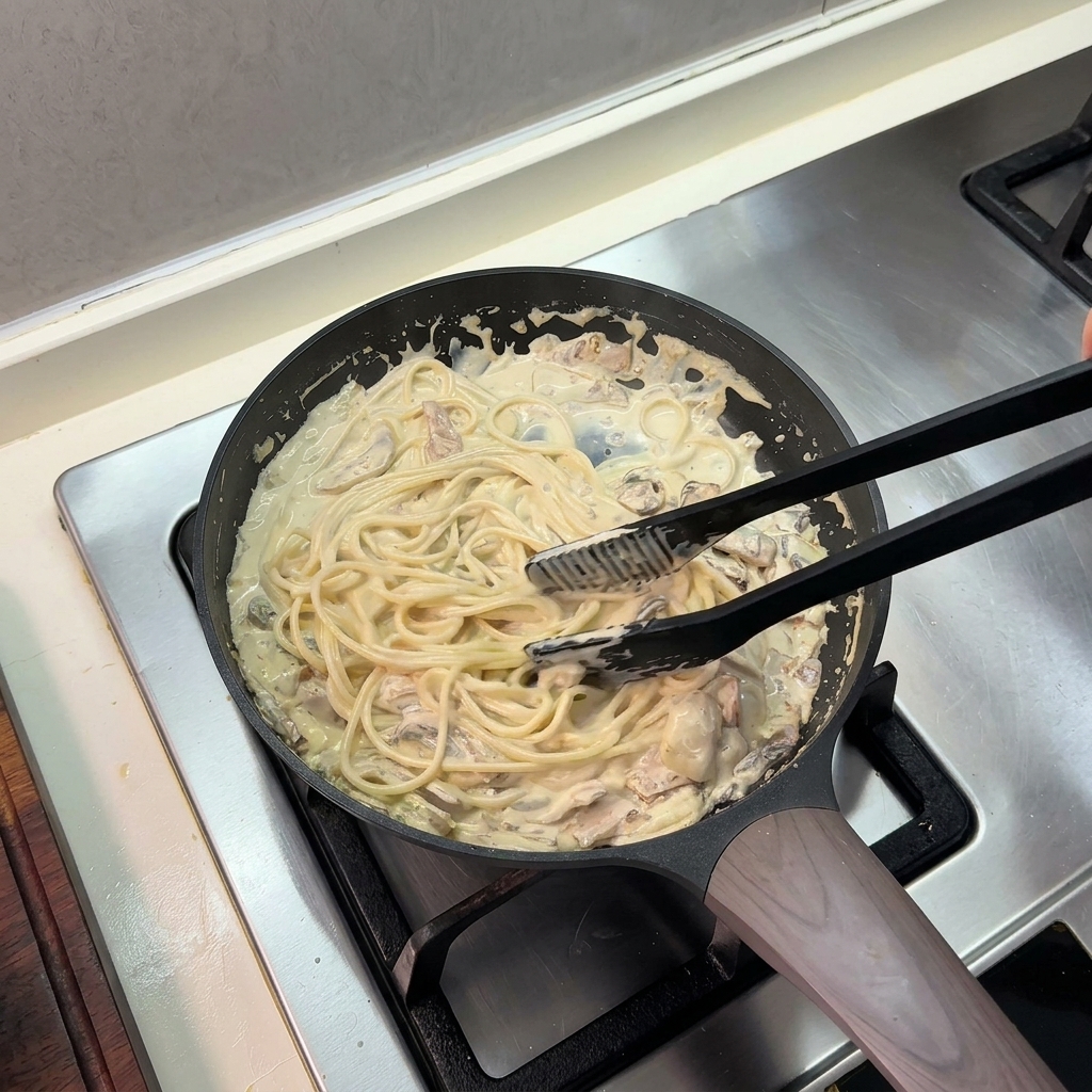 Czarne szczypce kuchenne mieszające spaghetti w gęstym sosie śmietanowym na patelni.