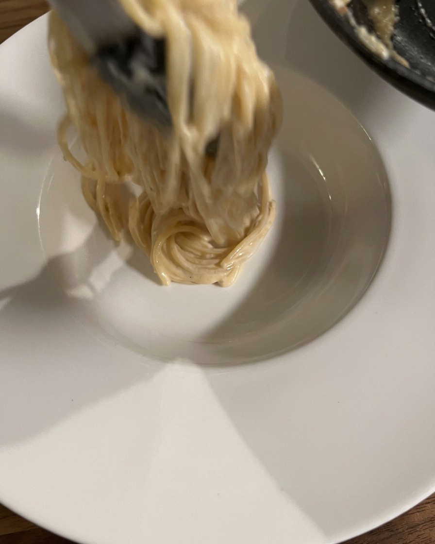 Szczypce unoszące i owijające porcję kremowego spaghetti nad czystą, białą, ceramiczną miską.