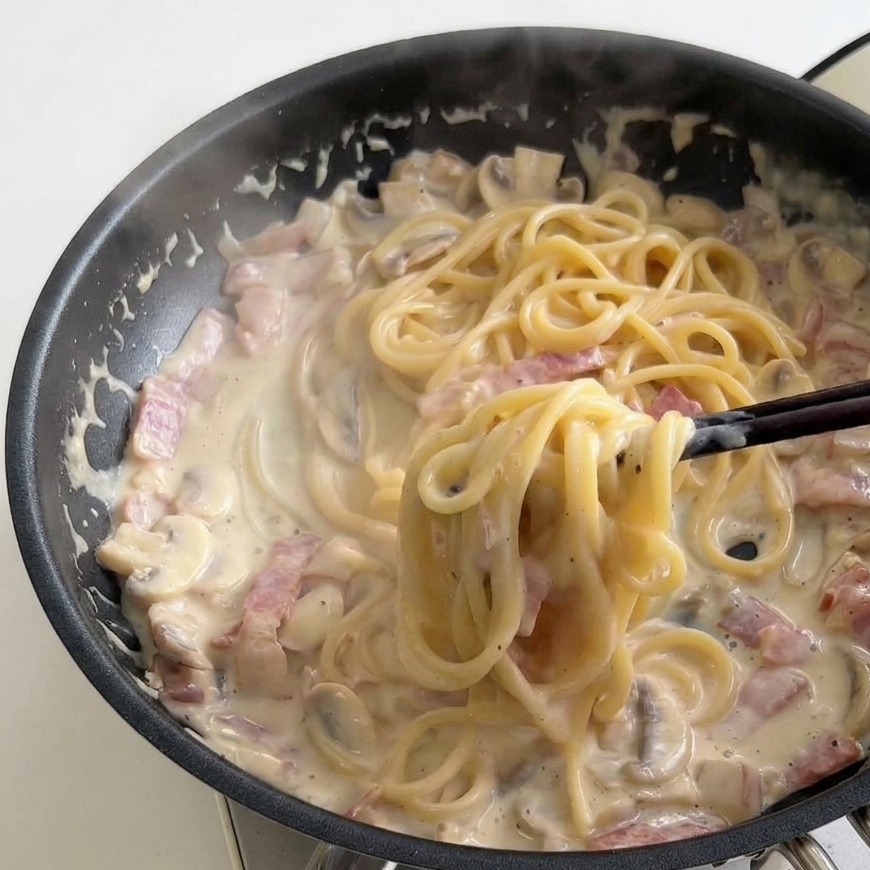 Pałeczki mieszające kremowe spaghetti na patelni w gęstniejącym, bulgoczącym sosie.