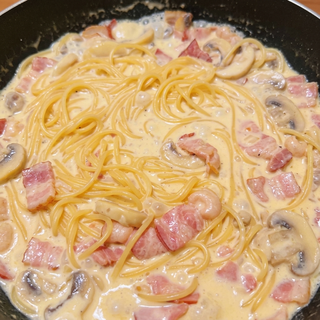 Ugotowany makaron spaghetti wymieszany z bogatym, kremowym sosem, boczkiem i pieczarkami na ciemnej patelni.