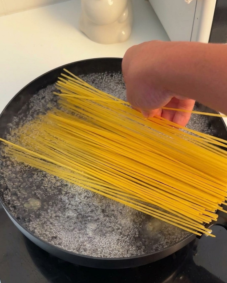 Dłoń wkładająca pęczek suchego makaronu spaghetti do czarnej patelni wypełnionej wrzątkiem.