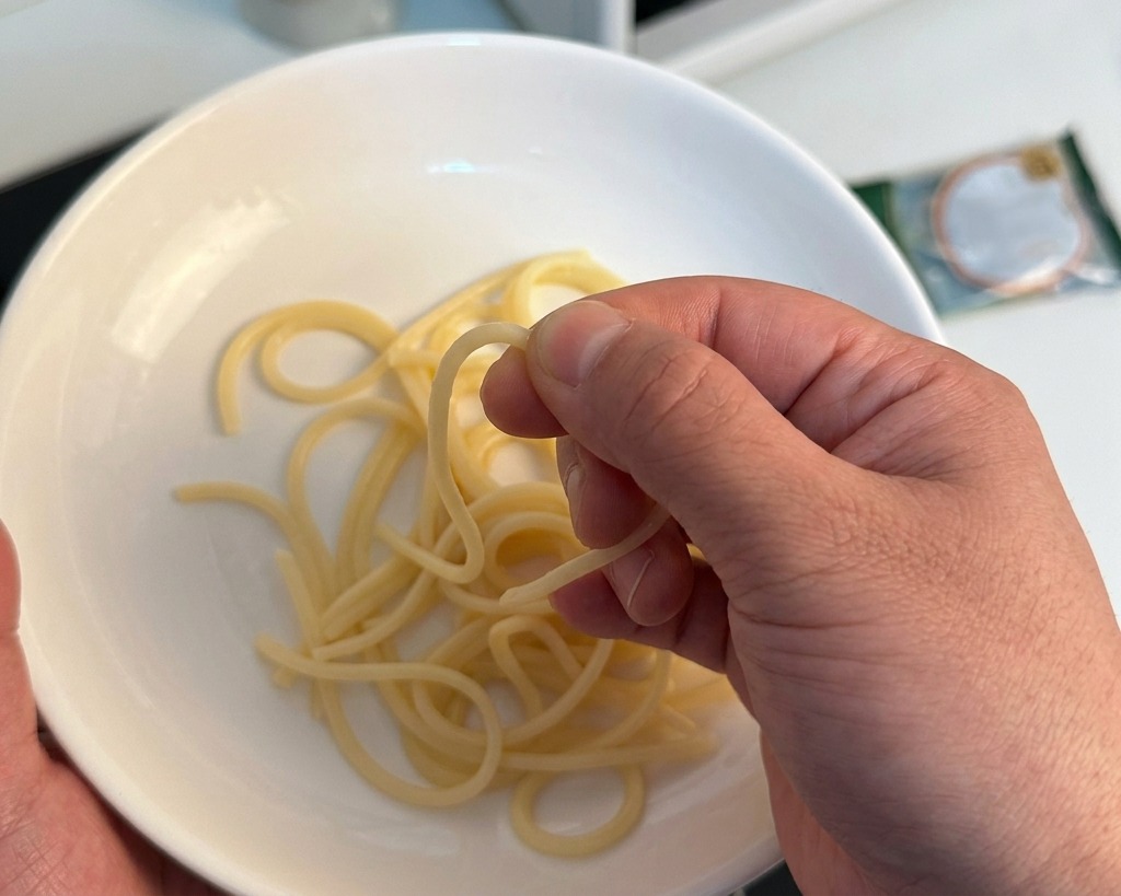 Dłoń sprawdzająca palcami sprężystość nitki spaghetti nad białą miską, aby ocenić czy jest gotowa.