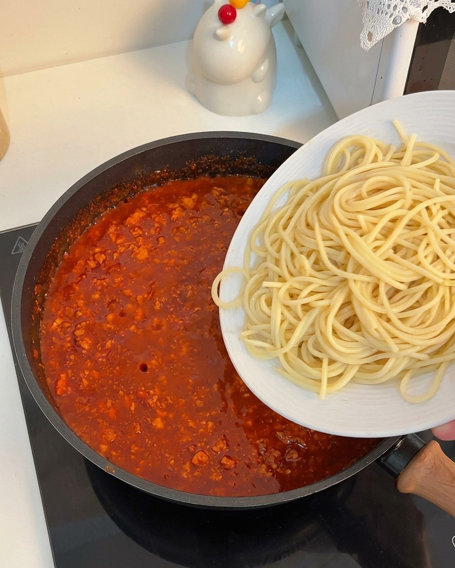 Ugotowany makaron spaghetti nakładany z białego talerza bezpośrednio do patelni z gęstym sosem.
