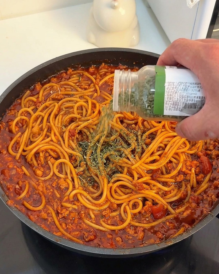 Dłoń posypująca suszoną natką pietruszki makaron spaghetti w gęstym, czerwonym sosie mięsnym.