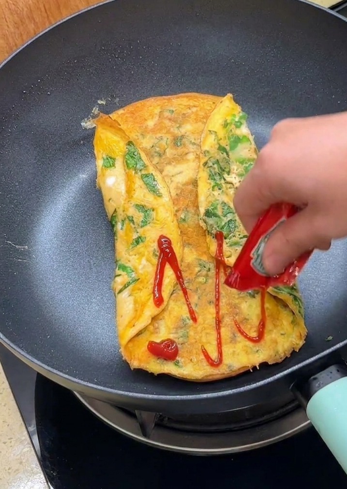 Ręka wyciskająca ketchup na powierzchnię zawiniętego omletu na patelni.
