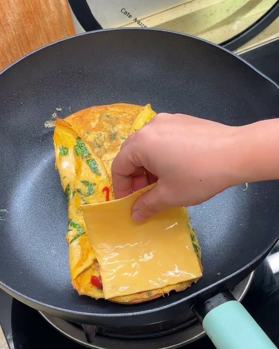 Ręka kładąca plaster żółtego sera na złożony omlet na patelni.