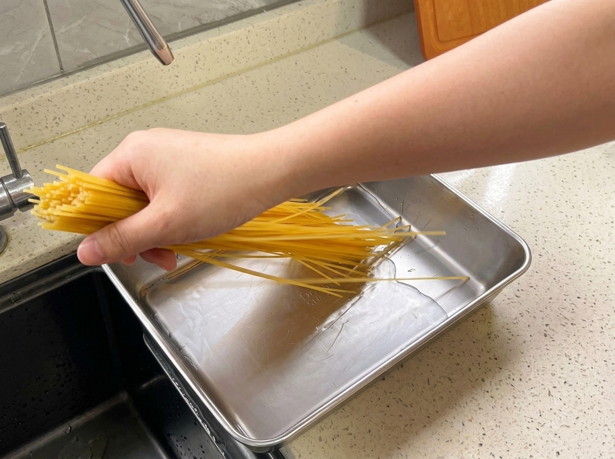 Ręka wkładająca pęczek żółtego spaghetti do prostokątnej metalowej tacy wypełnionej wodą na kuchennym blacie.