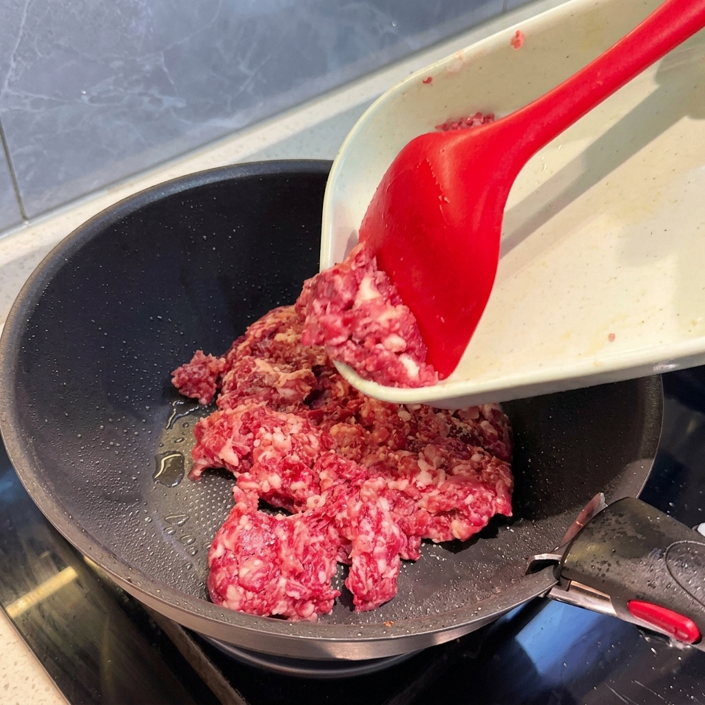 Surowe, grubo mielone mięso przesuwane z deski do krojenia na ciemną patelnię za pomocą czerwonej łopatki.