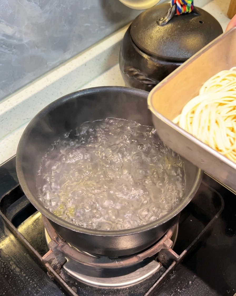 Wkładanie porcji długiego spaghetti z metalowej tacy do garnka z wrzącą wodą.