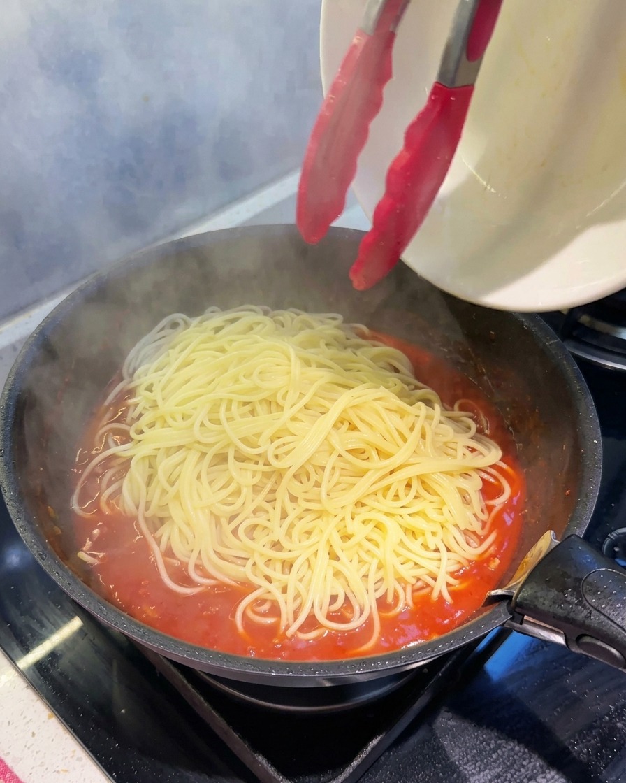 Szczypce przekładające dymiące, świeżo ugotowane spaghetti na patelnię pełną gęstego sosu mięsnego.