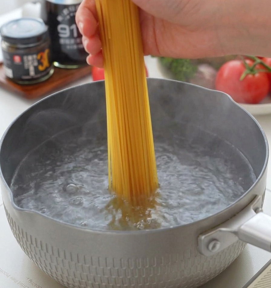 Ręka wkładająca wiązkę suchego spaghetti pionowo do szarego garnka z mocno wrzącą wodą.