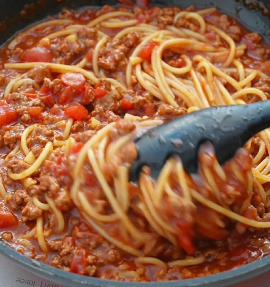 Czarne szczypce mieszające ugotowane spaghetti z gęstym sosem mięsnym na patelni.