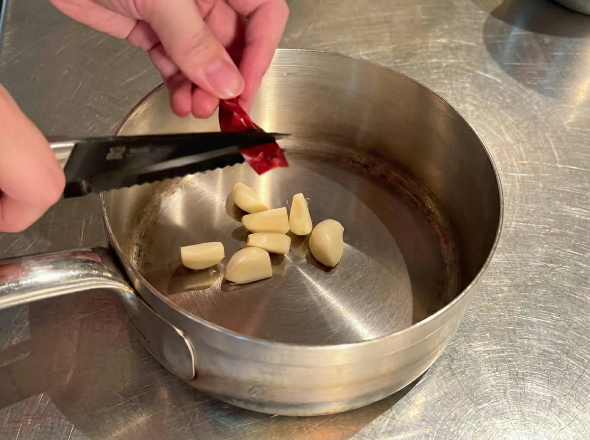 Dłonie tnące suszoną papryczkę chili nożyczkami nad garnkiem z czosnkiem.